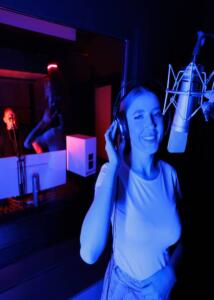 A young woman with headphones records a song into a studio microphone in a recording music studio barcelona against the background of a sound engineer behind glass